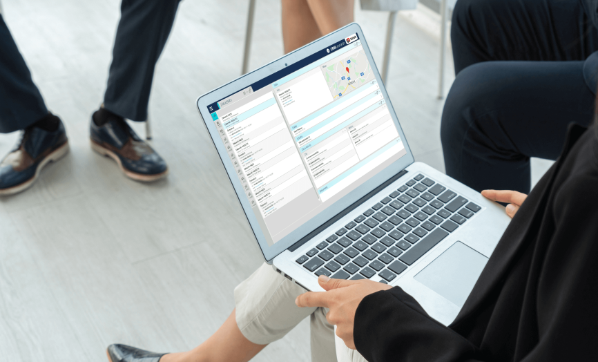 A woman sitting in a room holds an open laptop on her lap displaying an energy or CRM software interface with tables, a map view, and data lists; other people are seated nearby.