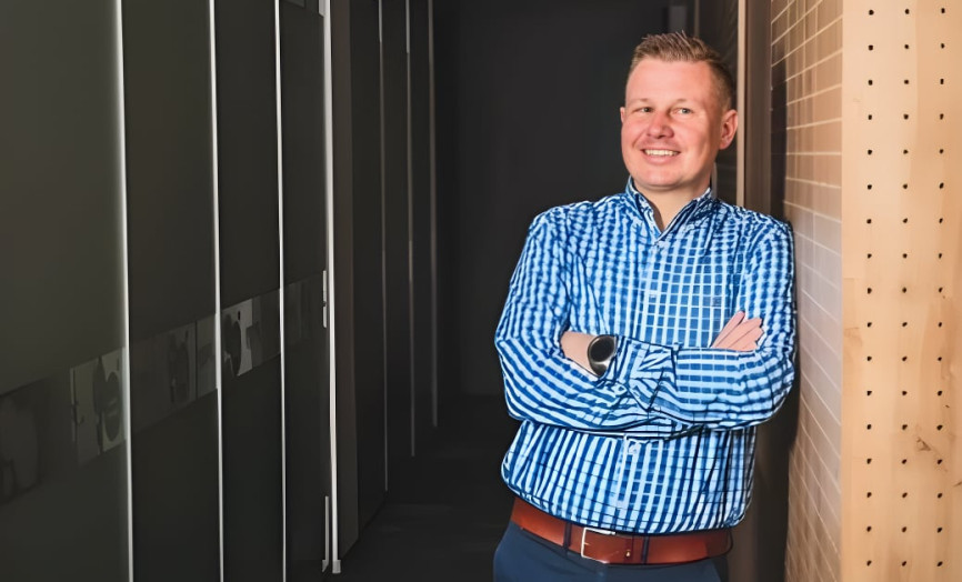 Muž v kostkované košili stojí v moderní chodbě kanceláře, usmívá se a má zkřížené ruce.